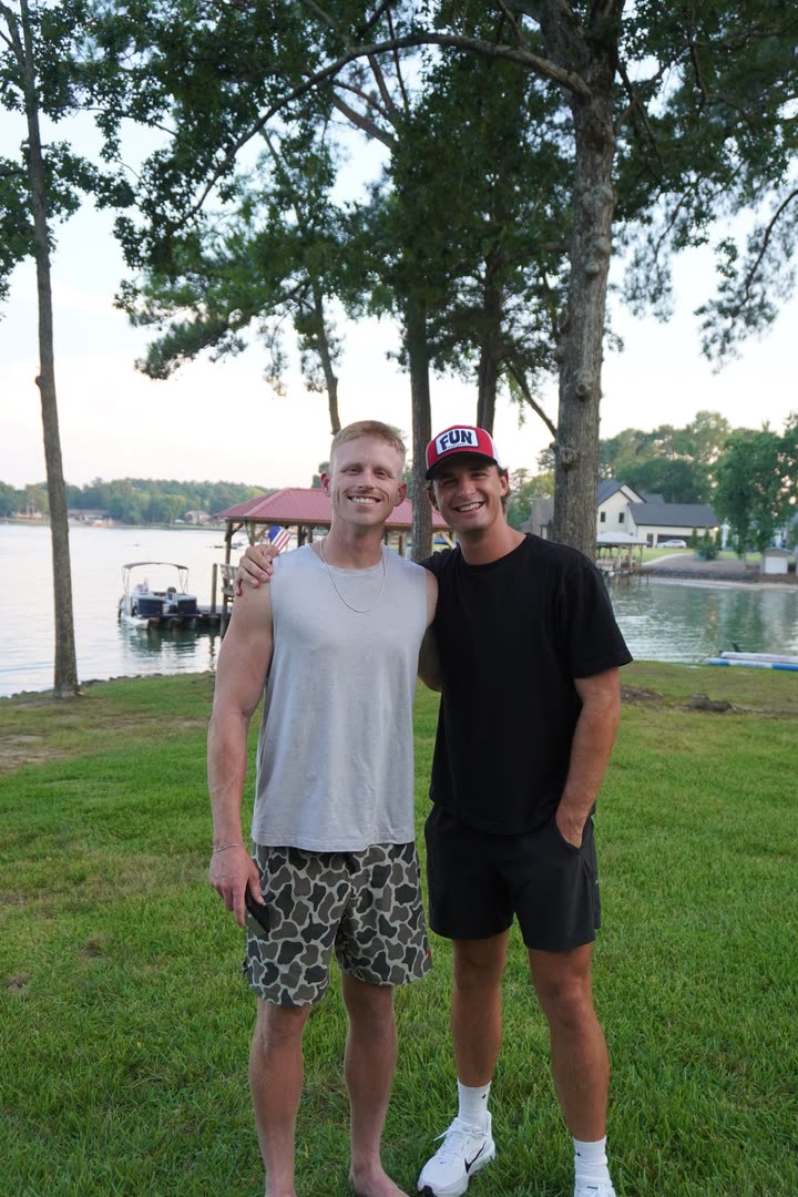 Zach von der Linden standing on a lawn beside another man, with a lake, a dock and trees behind them