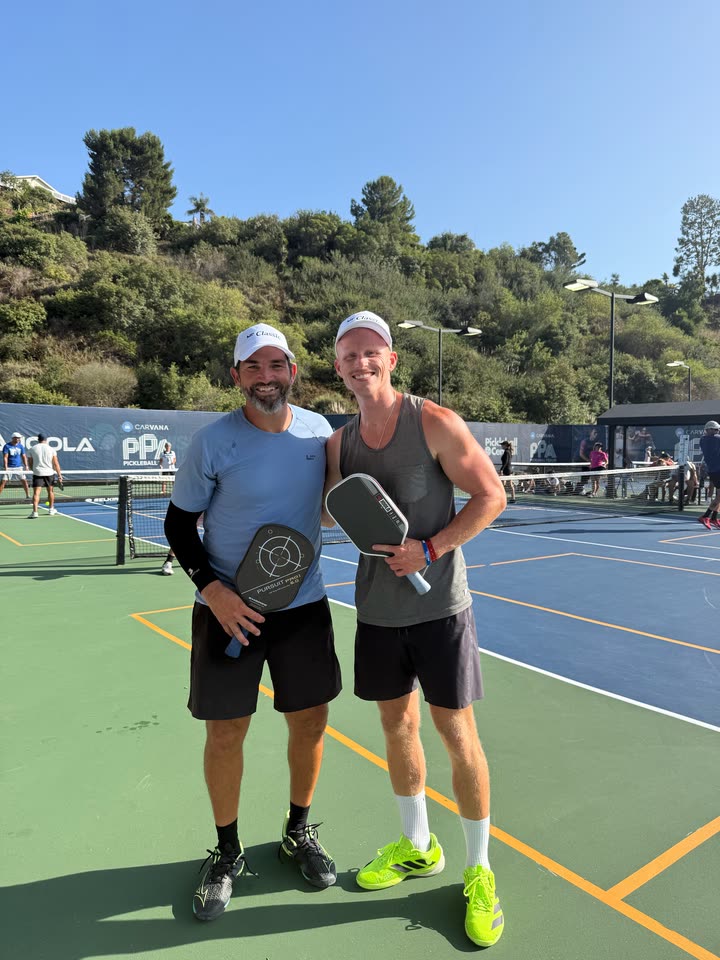 Zach von der Linden holding a pickleball paddle beside another man on an outdoor court, with tournament banners behind them