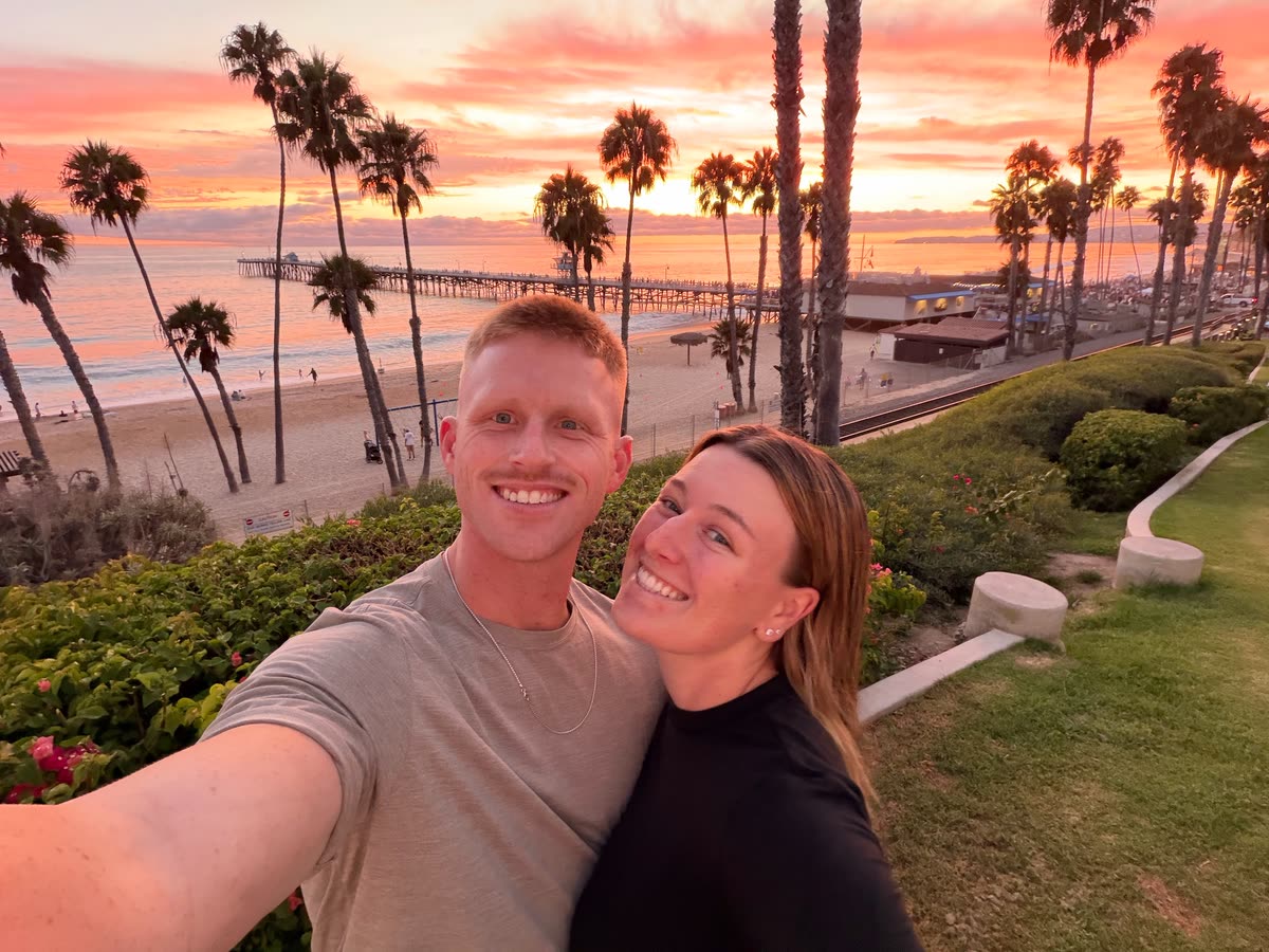 Zach von der Linden taking a selfie with a woman above a beach and pier at sunset, with palm trees behind them