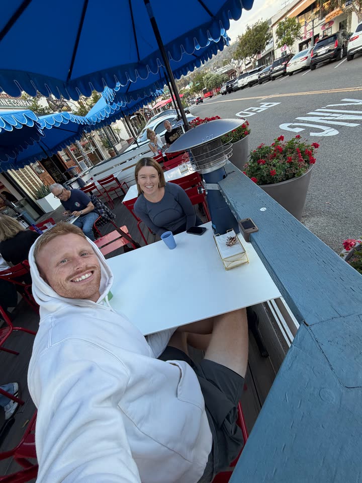 Zach von der Linden in a white hoodie taking a selfie at an outdoor patio table under blue umbrellas, with a woman seated at the table behind him
